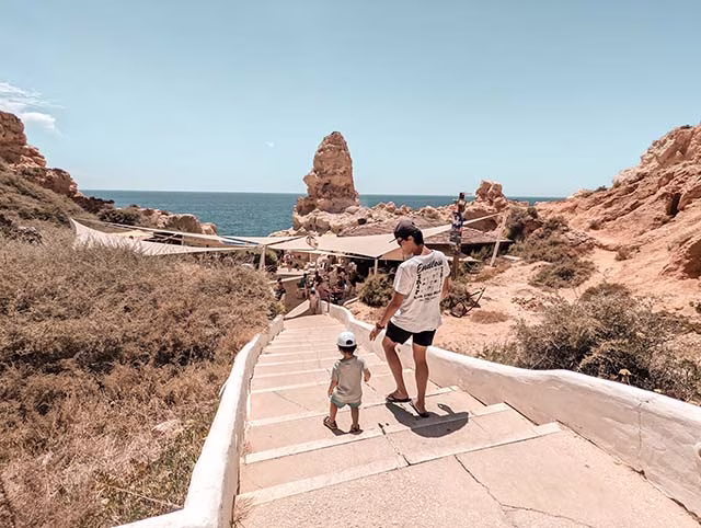 a delightful moment of a millennial family during their travels in Algarve, Portugal. The family, consisting of two adults and two children, are seen from behind, standing on a sandy beach and looking out towards the ocean. The sky is a brilliant blue, and the waves gently lap onto the shore, creating a serene and picturesque setting. The family's sense of wonder and adventure is palpable, encapsulating the joy of minimalist family travel.