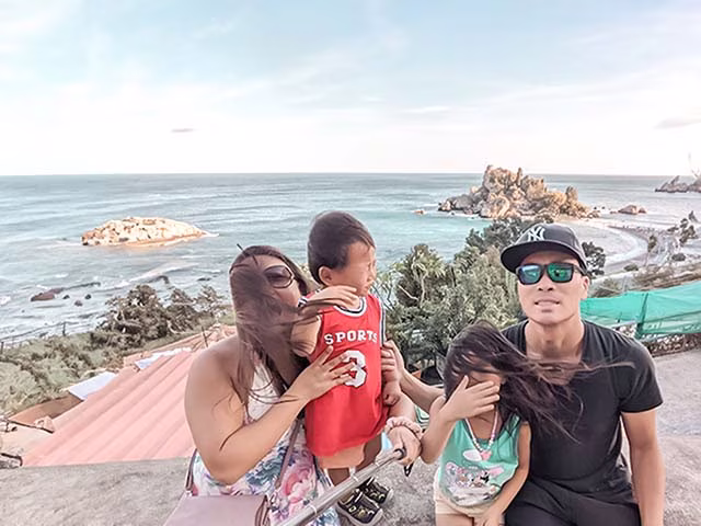 a humorous moment of a millennial family during their travels around the world. The family, consisting of two adults and two children, are seen in a selfie, but it's not your typical picture-perfect shot. Instead, it's a candid moment showing real-life family dynamics, with the children making funny faces and the parents laughing. This image beautifully encapsulates the joy, challenges, and unpredictability of family travel.