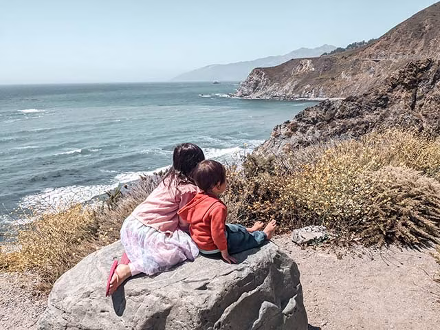 a family on a road trip along the California Coast. The family, composed of two adults and two children, are seen from behind, standing on a sandy beach and looking out towards the ocean. The sky is a vibrant blue, and the waves crash onto the shore, creating a beautiful and tranquil scene. The family appears to be enjoying the view, embodying the spirit of adventure and the joy of travel.