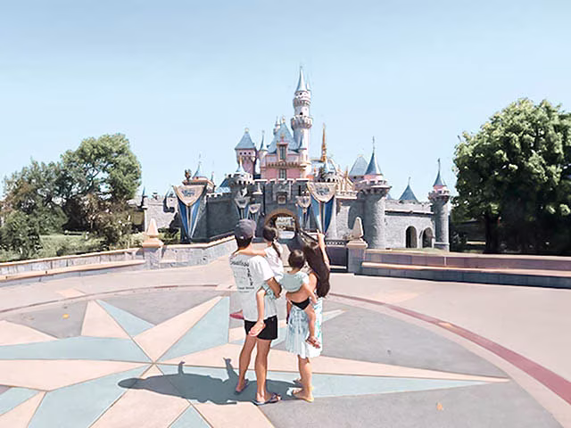 a joyful moment of a millennial family at Disneyland in Anaheim, California. The family, consisting of two adults and two children, are seen from behind, walking towards the iconic Disneyland castle. The castle, bathed in warm, golden light, stands majestically against a clear blue sky. The family's excitement and anticipation are palpable, embodying the magic and wonder of a Disneyland adventure.