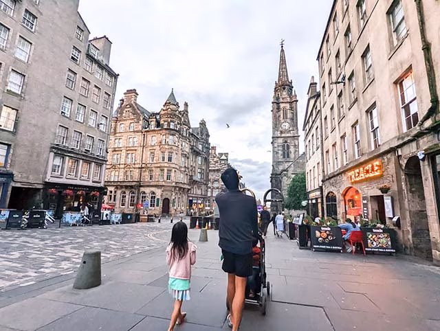 a millennial family on their travels in Edinburgh. The family, consisting of two adults and two children, are seen from behind, looking out over a panoramic view of the city. The skyline of Edinburgh, with its historic buildings and landmarks, stretches out before them under a sky filled with soft, diffused light. The family appears to be in a moment of awe, taking in the beauty and grandeur of the city. This image beautifully captures the essence of family travel and the joy of exploring new places together.
