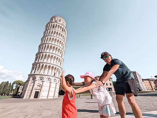 a millennial family on their travels in Pisa, Italy. The family, consisting of two adults and two children, are seen from behind, looking towards the iconic Leaning Tower of Pisa. The tower, with its distinctive tilt, stands against a clear blue sky. The family's sense of wonder and adventure is evident, capturing the essence of their journey around the world.