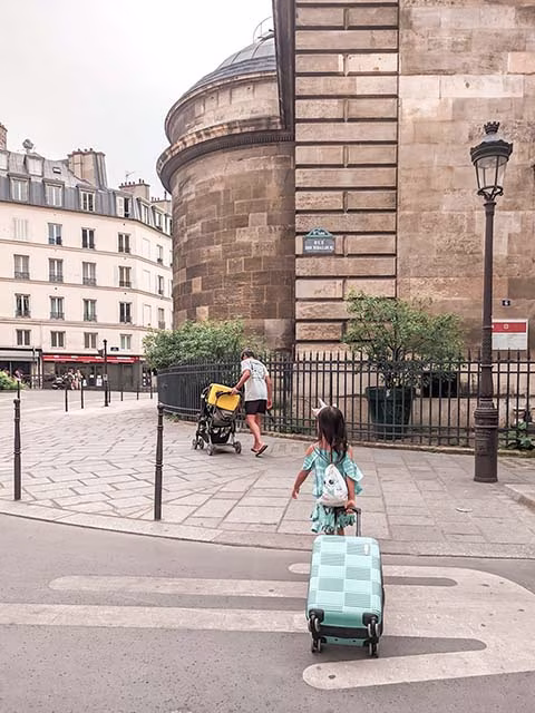 a millennial family on their minimalist travel journey around the world. The family, consisting of two adults and two children, are seen from behind, walking down a path with their light carry-on luggage. The path is lined with trees, creating a serene and peaceful setting. The family's minimalist approach to travel is evident in their light luggage, embodying the spirit of simplicity and freedom that comes with minimalist travel.