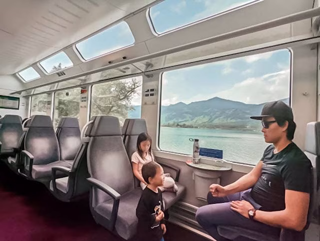 a millennial family on a train journey through the picturesque landscapes of Switzerland. The family, consisting of two adults and two children, are seen from behind, looking out of the train window at the stunning scenery. The view outside is a breathtaking panorama of lush green fields, rolling hills, and distant mountains. The family's sense of awe and wonder is palpable, encapsulating the joy and adventure of minimalist travel.