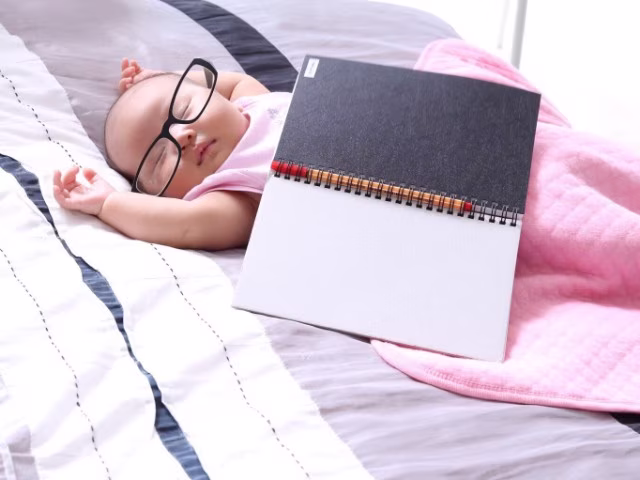 cute baby sleeping wearing oversized glasses with arms stretched out, diary next to her covered with pink blanket
