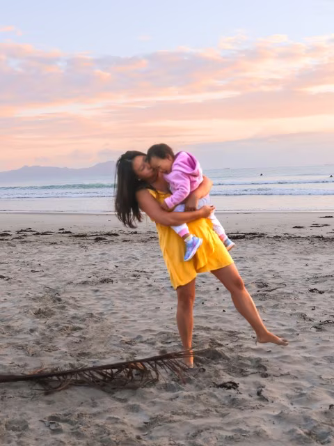 Mother wearing a yellow dress, playfully holding a baby on the beach in golden hour