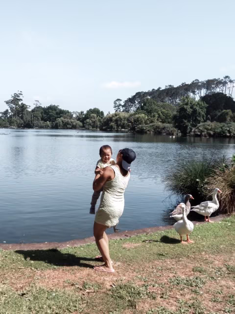 Mother holding up a baby with bent knee in Western Spring Auckland New Zealand by the lake. Postpartum back pain self care